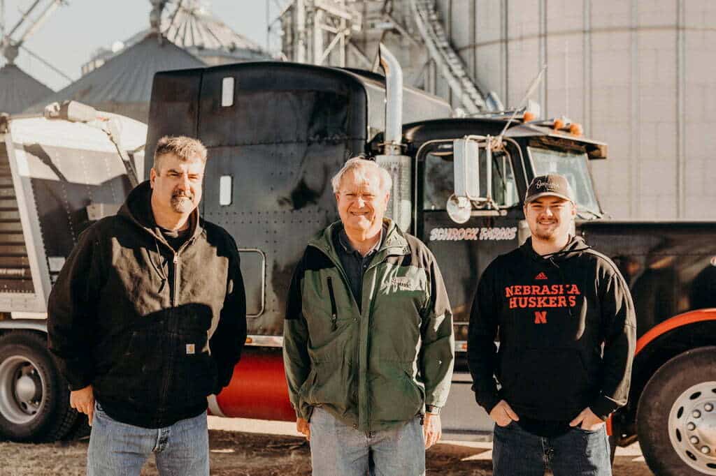 Three generations of farmers standing in front of a semi and grain bins