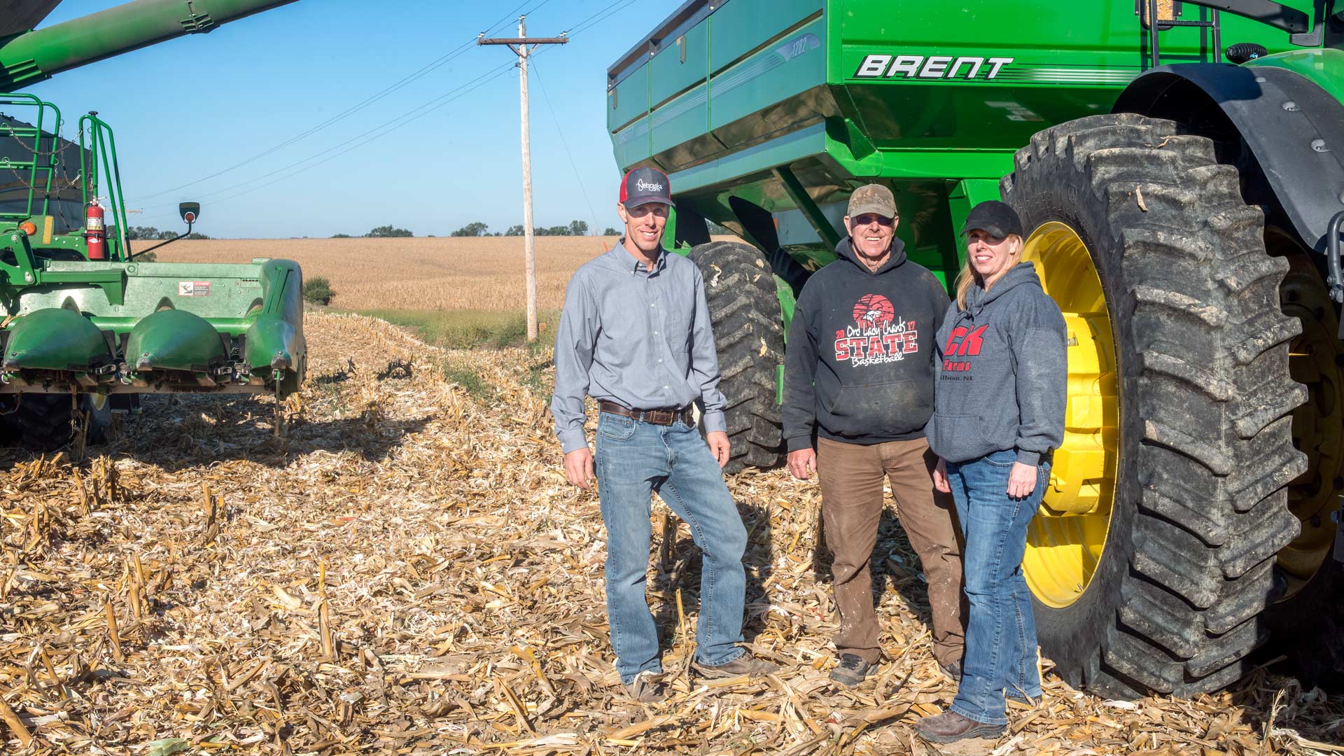 Three farmers stand next to farm equipment in a cornfield after harvest is complete.