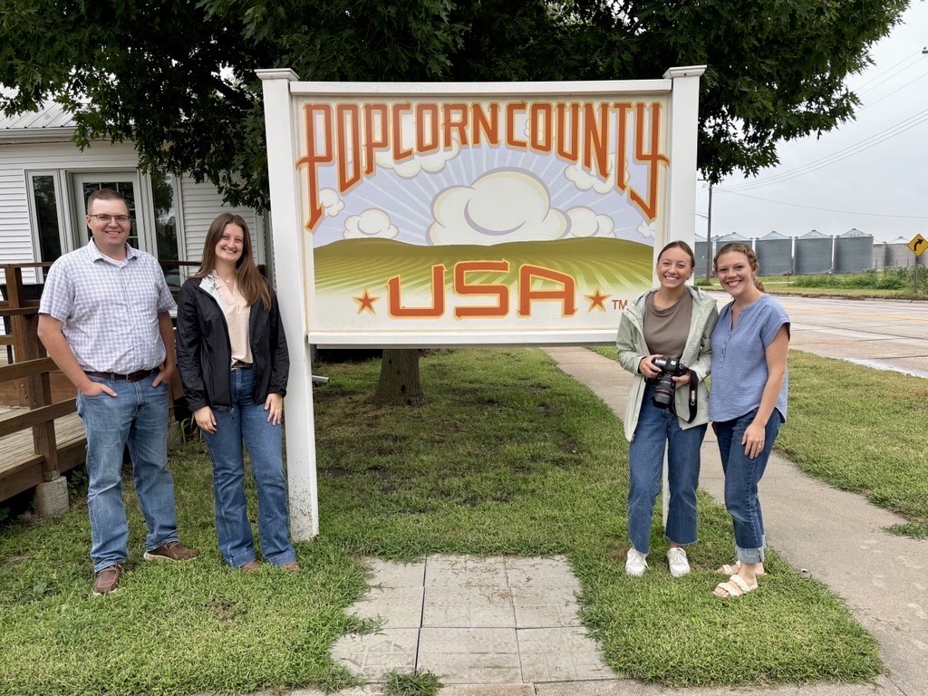 Four people pose outdoors next to a large sign that reads "Popcorn County USA".