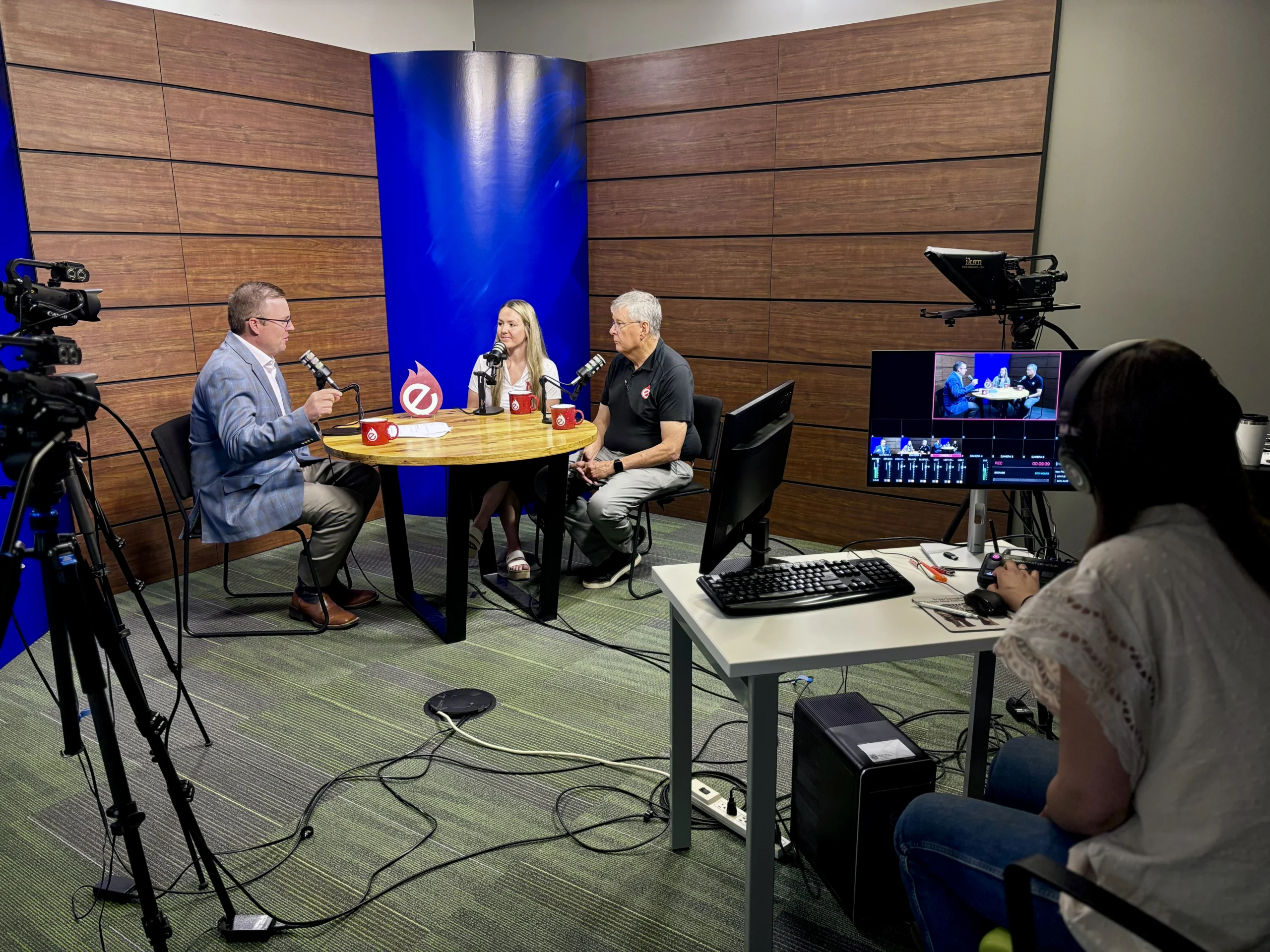 Rural Radio Network- Lincoln, NE - Three people speak around a circular table in a studio while a production operator monitors the recording from a control desk.