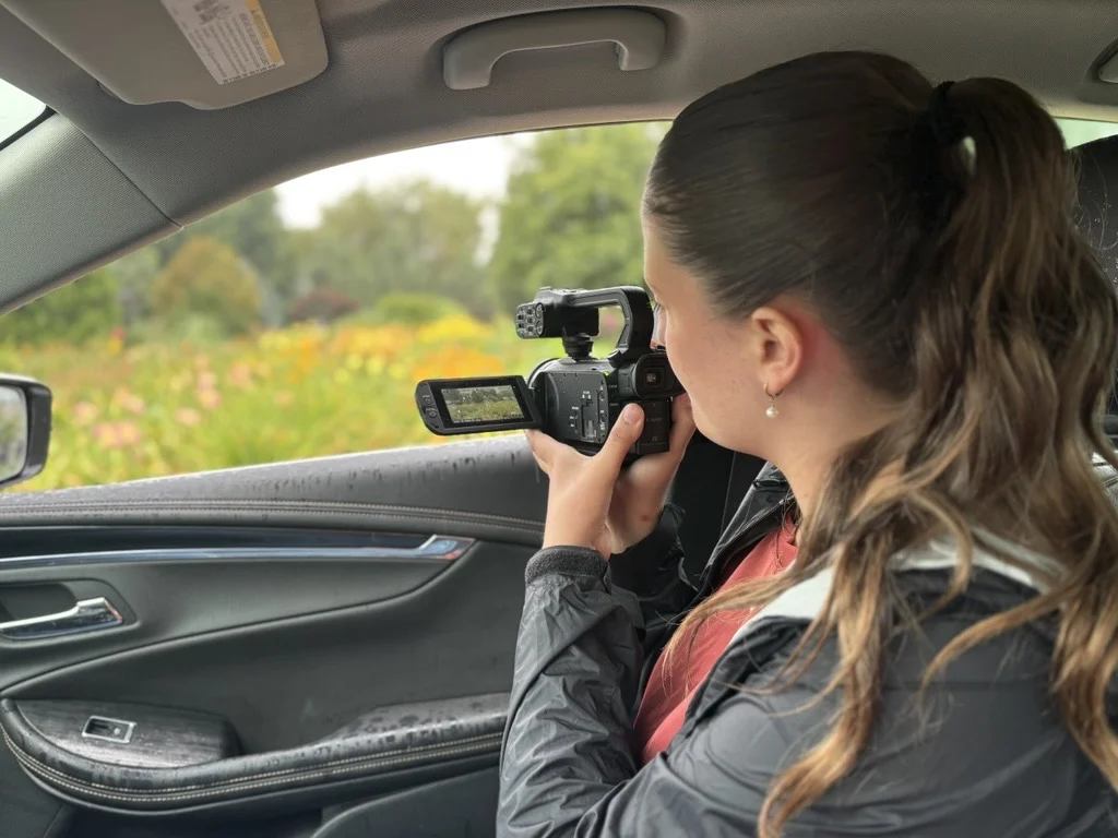 A woman holding a video camera looks through the viewfinder while filming out of an open car window.